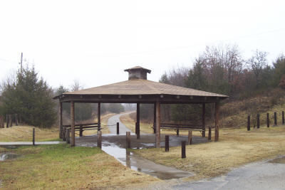 A wooden gazebo on a misty day, surrounded by a grassy area and a winding road in the background.