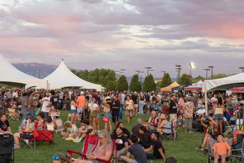 A lively outdoor festival scene with large crowds, tents, and a colorful sunset in the background.