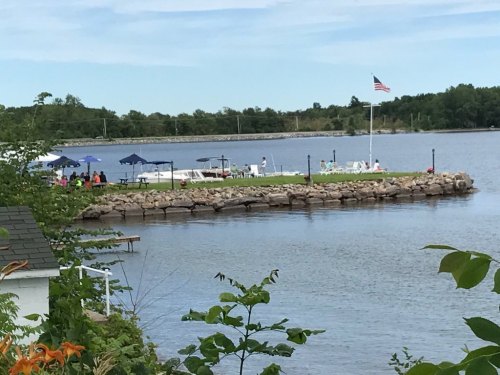 A serene lakeside scene with a grassy area, umbrellas, and an American flag, surrounded by calm water and greenery.