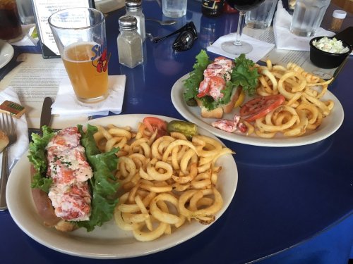 Two plates of lobster rolls with curly fries, a glass of beer, and a glass of red wine on a blue table.