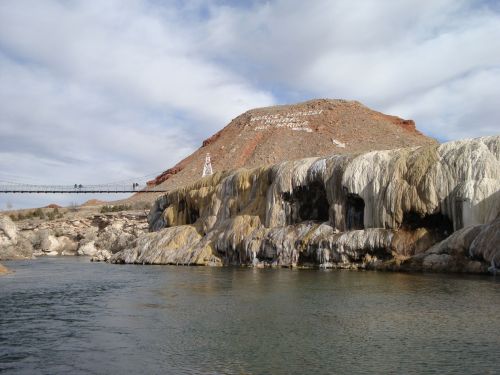 A scenic view of a rocky hill with a bridge, overlooking a river with mineral formations and cloudy skies.