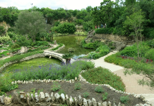 A serene garden landscape featuring a pond, stone pathways, and lush greenery under a cloudy sky.