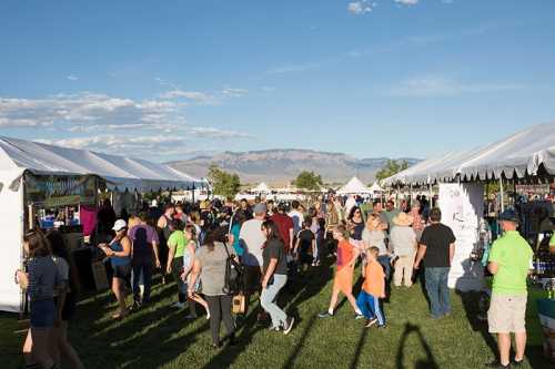 A lively outdoor festival with crowds of people browsing tents against a backdrop of mountains and blue sky.