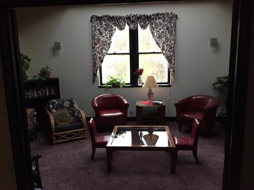 Cozy sitting area with two red chairs, a glass coffee table, and plants, illuminated by natural light through a window.