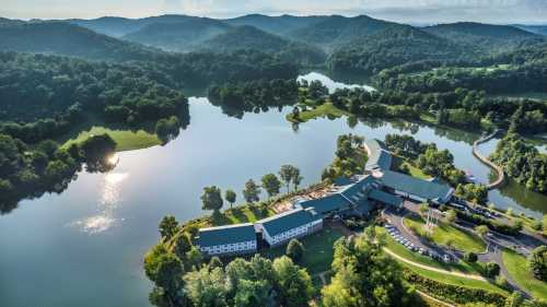 Aerial view of a serene lakeside resort surrounded by lush green hills and trees, reflecting in the calm water.