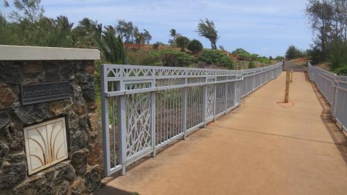 A metal bridge with decorative railings spans a pathway surrounded by greenery under a cloudy sky.