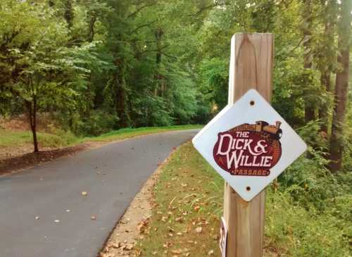 Sign for the Dick & Willie Passage along a tree-lined, paved path in a wooded area.