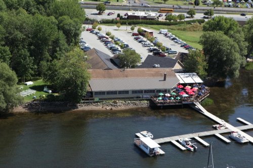 Aerial view of a lakeside restaurant with a dock, surrounded by trees and a parking lot, on a sunny day.