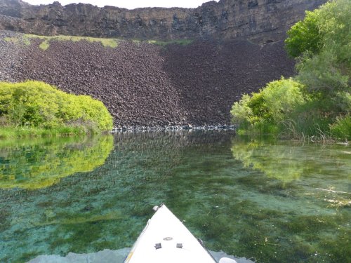 A kayak on clear water reflects a rocky cliff and lush greenery in a serene natural setting.