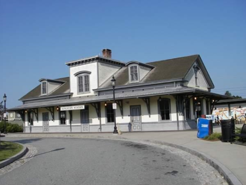 Historic train station with a white and gray exterior, featuring a sloped roof and surrounding paved area.