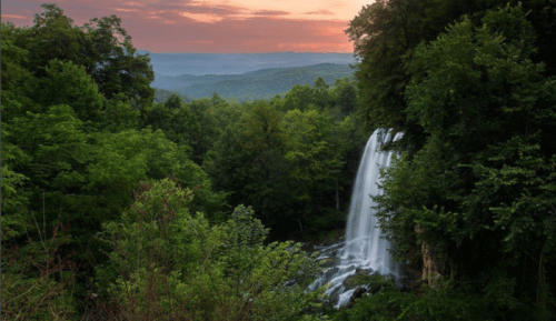 A serene waterfall cascades through lush green trees at sunset, with distant mountains visible in the background.