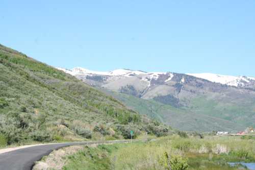 A scenic view of a winding road surrounded by green hills and snow-capped mountains under a clear blue sky.