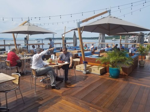 Outdoor dining area with umbrellas, people enjoying meals, and a waterfront view. Relaxed atmosphere with wooden decking.