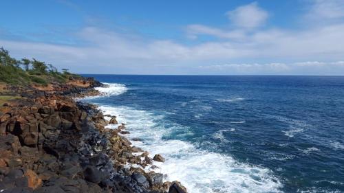 A rocky coastline meets a vibrant blue ocean under a partly cloudy sky. Waves crash against the shore.