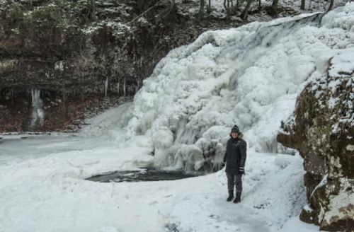 A person stands near a frozen waterfall surrounded by snow-covered rocks and trees.