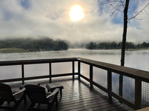 A serene lakeside view with mist rising, sun shining through clouds, and two chairs on a wooden deck.