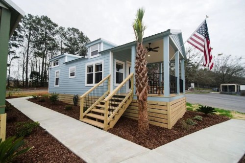 A blue house with a porch, palm tree, and American flag, surrounded by landscaped pathways and greenery.
