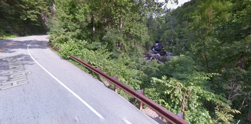 A winding road alongside a lush green forest and a rocky stream.