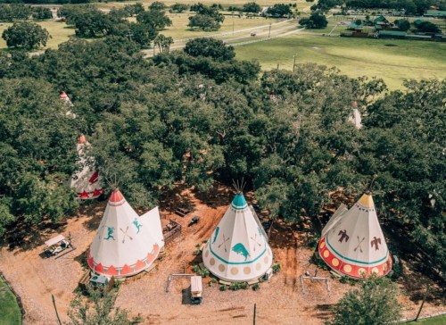 Aerial view of colorful teepees nestled among trees, with a grassy area and road in the background.