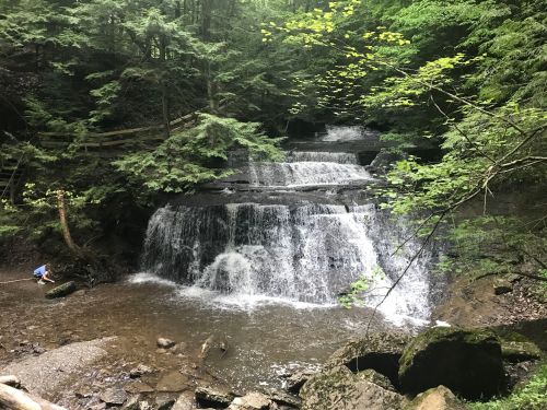 A serene waterfall cascades over rocks in a lush green forest, with a person exploring the water's edge.