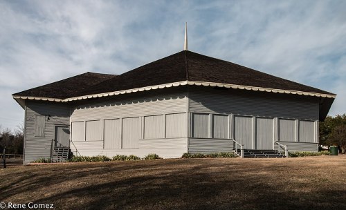 A gray wooden building with a peaked roof, featuring boarded windows and a grassy area in front.
