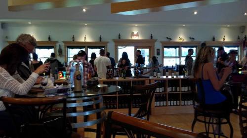 A busy wine tasting room with people socializing, seated at tables, and a display of wine bottles in the background.