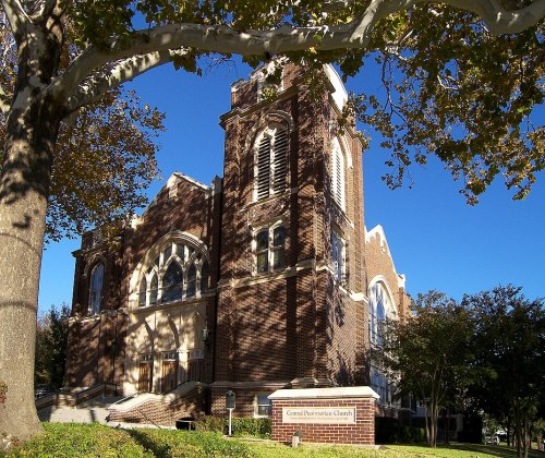 A brick church building with a tall steeple, surrounded by trees and a clear blue sky.