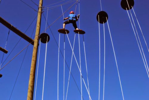 A person in a blue shirt and helmet climbs a tall structure with circular platforms against a clear blue sky.