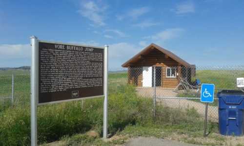 Sign for Vore Buffalo Jump with a small building nearby, set against a clear blue sky and grassy landscape.