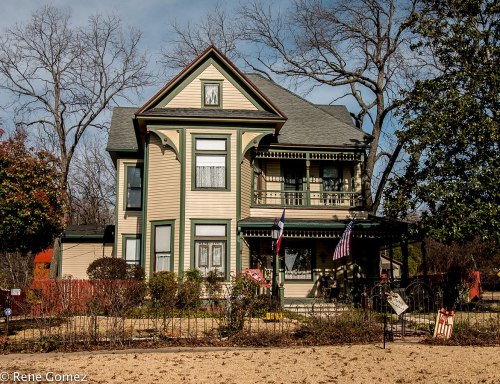A charming Victorian-style house with green trim, surrounded by trees and a small garden, featuring American flags.