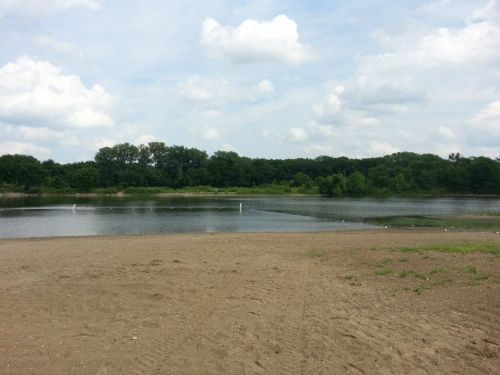 A serene lake surrounded by trees, with a sandy shore and cloudy sky above.