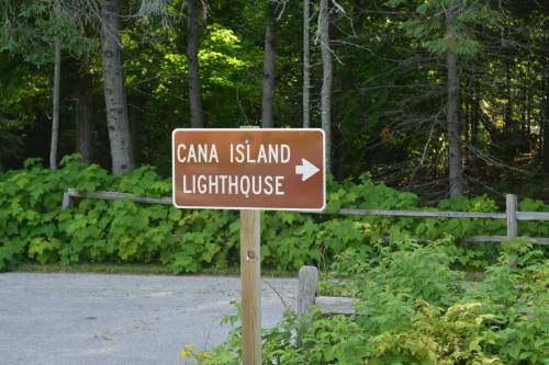 Brown sign pointing to Cana Island Lighthouse, surrounded by greenery and trees.