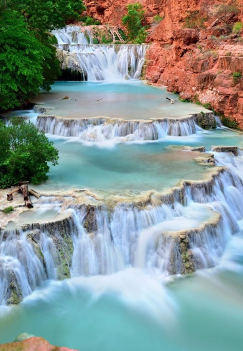 A serene waterfall cascades over rocky tiers into turquoise pools, surrounded by lush greenery and red rock formations.