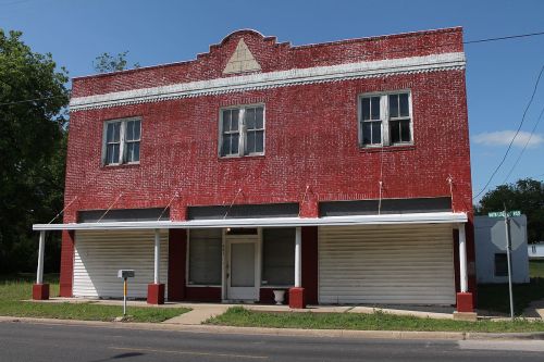 Abandoned red brick building with white trim, featuring boarded windows and a grassy area in front.