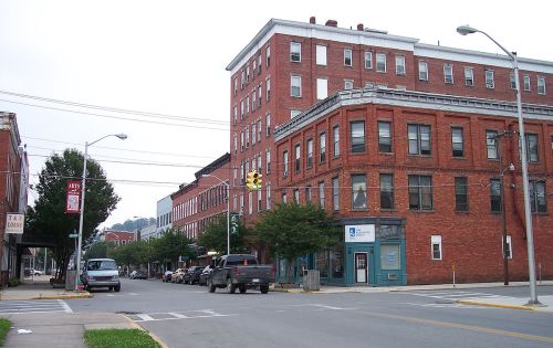 A street view of a small town featuring brick buildings, trees, and parked cars under a cloudy sky.