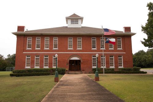 Historic red brick school building with American and Texas flags, surrounded by green grass and trees.