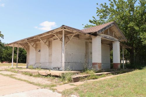 Abandoned building with a porch, overgrown grass, and trees nearby under a clear blue sky.