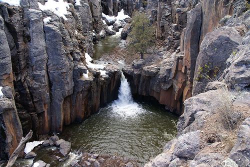 A waterfall cascades into a rocky canyon, surrounded by steep cliffs and patches of snow.