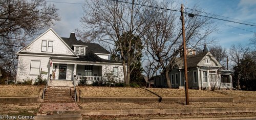 Two historic houses with porches, set on a grassy lot, under a cloudy sky. A power line runs along the street.