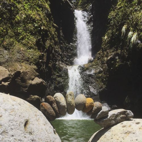 A serene waterfall cascades between rocky cliffs, with a stone arch formed by pebbles in the foreground.