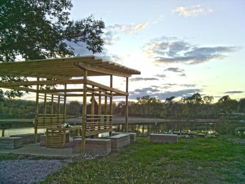 A wooden pavilion by a calm lake at sunset, surrounded by trees and a grassy area.