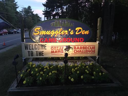 Sign for Smuggler's Den Campground, featuring "Welcome to the Great Maine Barbecue Challenge" with flowers below.