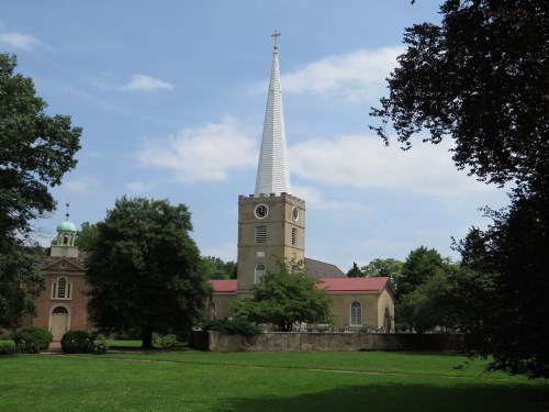 A church with a tall spire surrounded by trees and a grassy area under a blue sky with clouds.