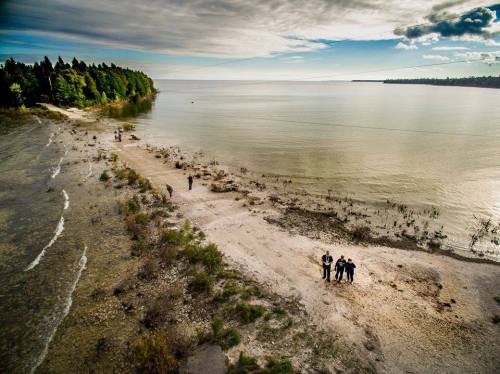 Aerial view of a sandy beach with people walking along the shore, surrounded by trees and calm water under a cloudy sky.