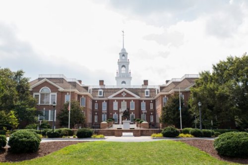 A brick building with a clock tower, surrounded by greenery and a cloudy sky.