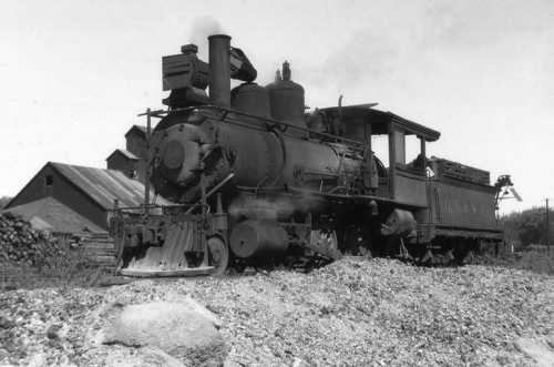 A vintage steam locomotive on a gravel track, with a barn and trees in the background, captured in black and white.