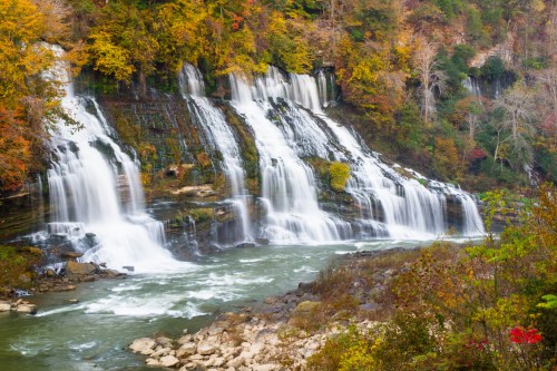 A picturesque waterfall cascading over rocks, surrounded by vibrant autumn foliage and a serene river below.