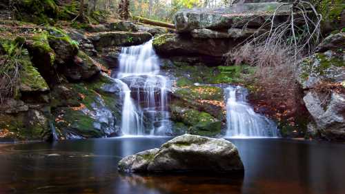 A serene waterfall cascades over moss-covered rocks into a calm pool, surrounded by autumn foliage.