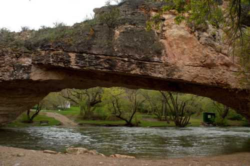 A natural stone arch over a river, surrounded by trees and greenery, with a pathway visible in the background.