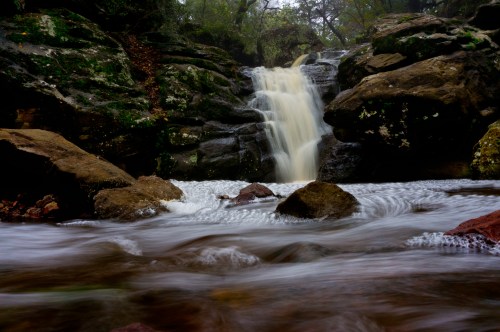 A serene waterfall cascades over rocks, surrounded by lush greenery and flowing water in a tranquil natural setting.